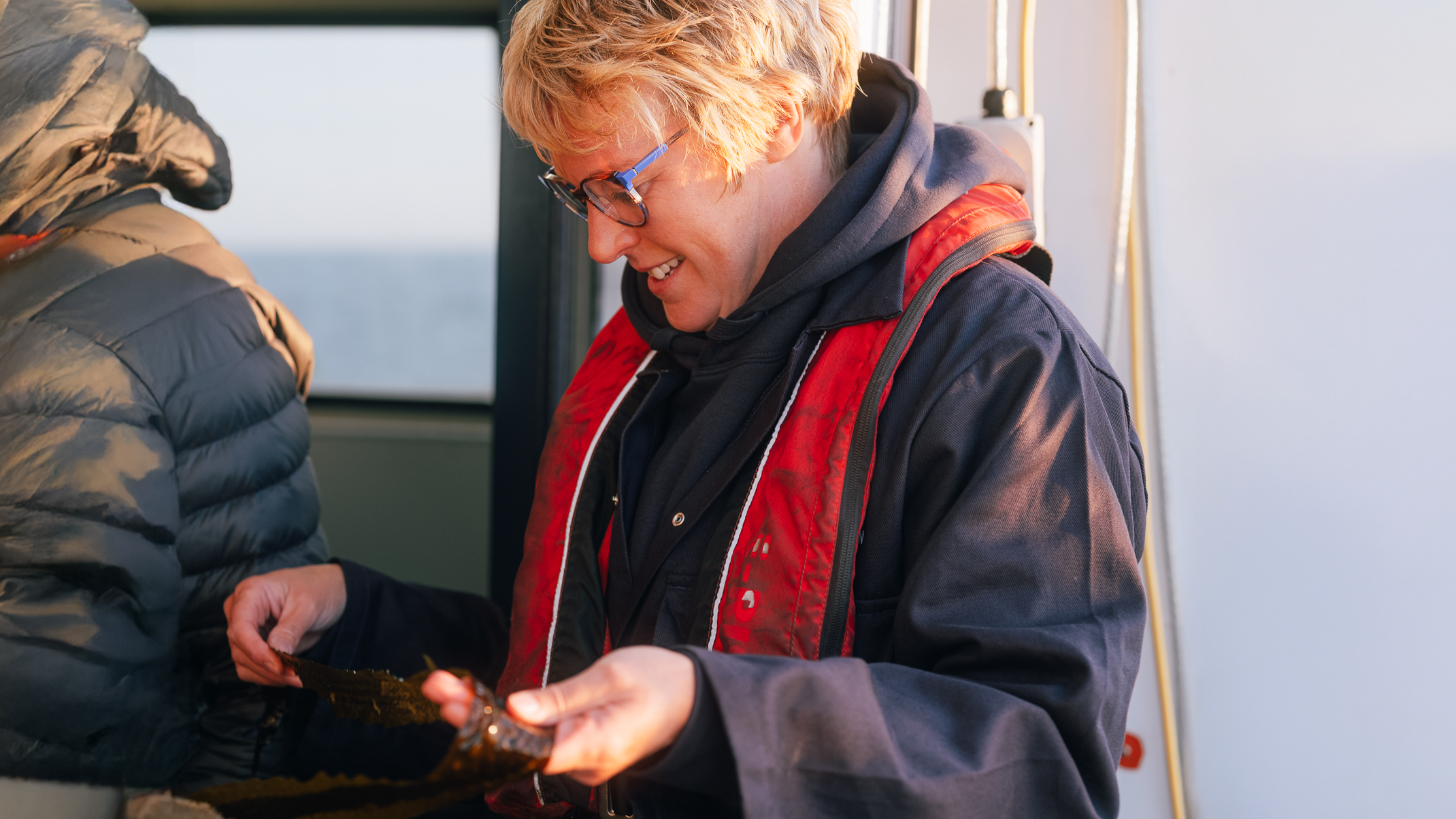 SeaGrown's General Manager, Jo Holborn, smiling in warm sunlight as she inspects a piece of freshly cultivated kelp on the deck of a boat at sea.