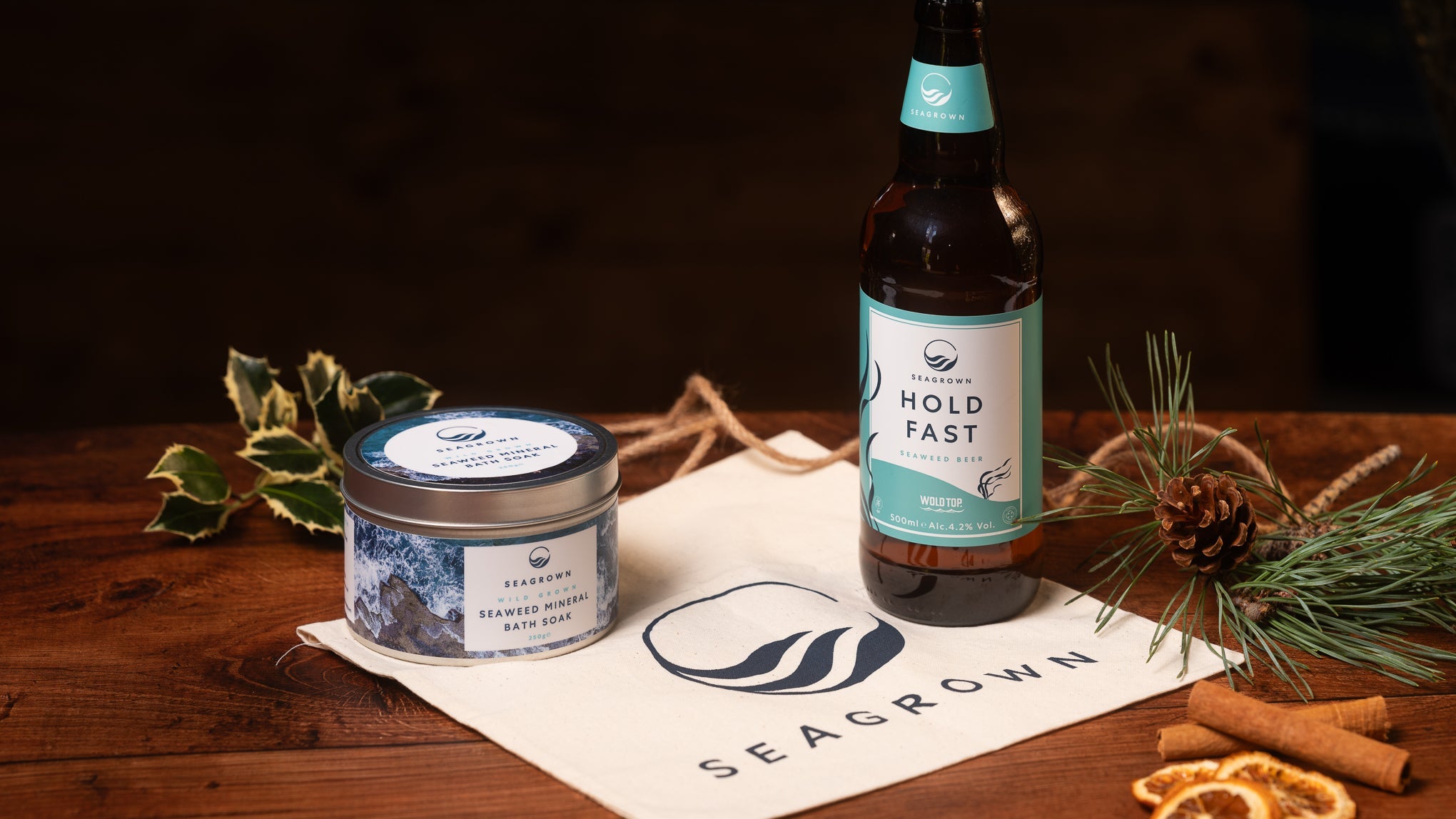 A tin of SeaGrown Seaweed Mineral Bath Soak and a bottle of Hold Fast seaweed beer on a SeaGrown cotton gift bag, with holly, pine, cinnamon and dried orange