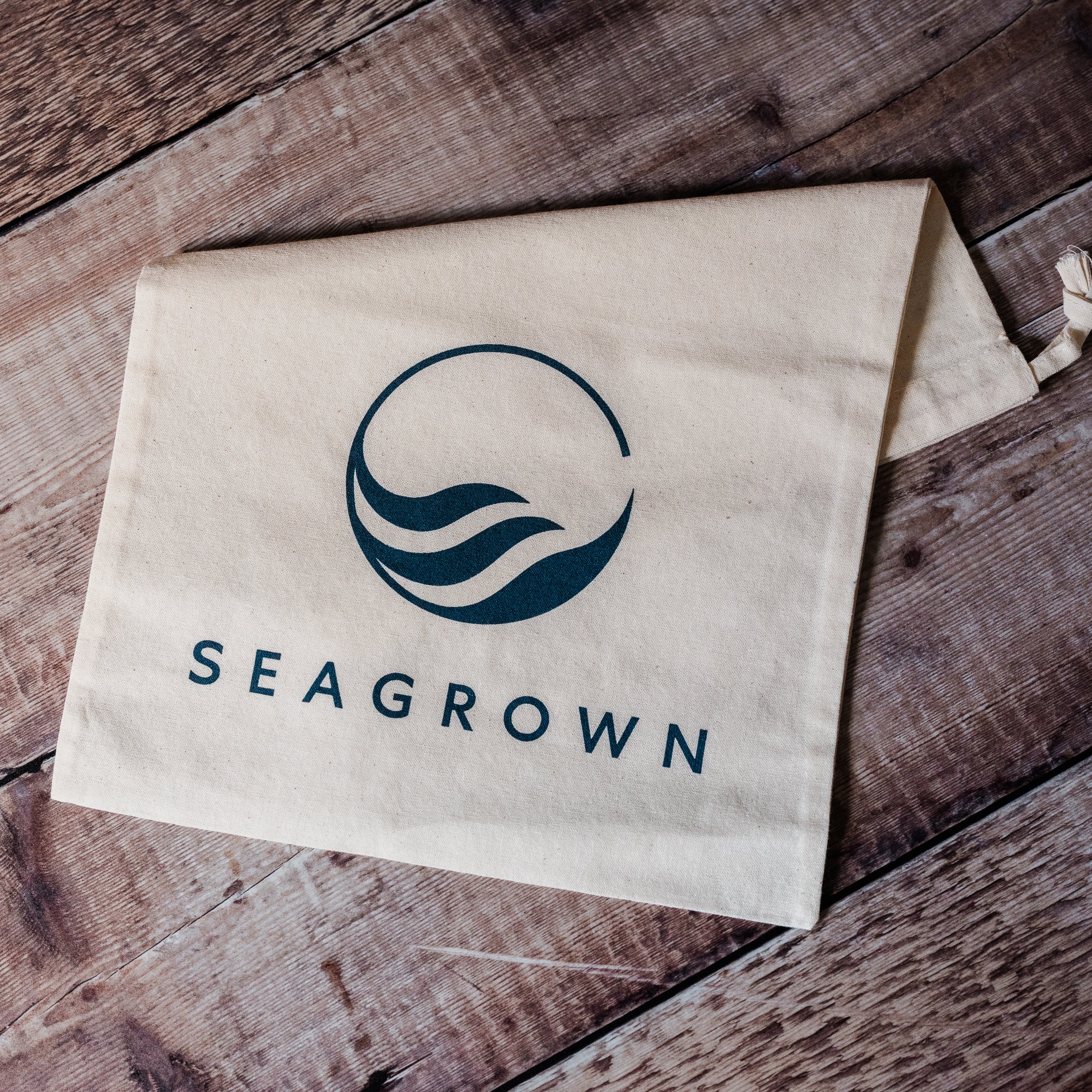 A beige canvas drawstring bag with a dark teal printed logo and the brand name 'SEAGROWN' laid flat on a rustic dark wood surface.